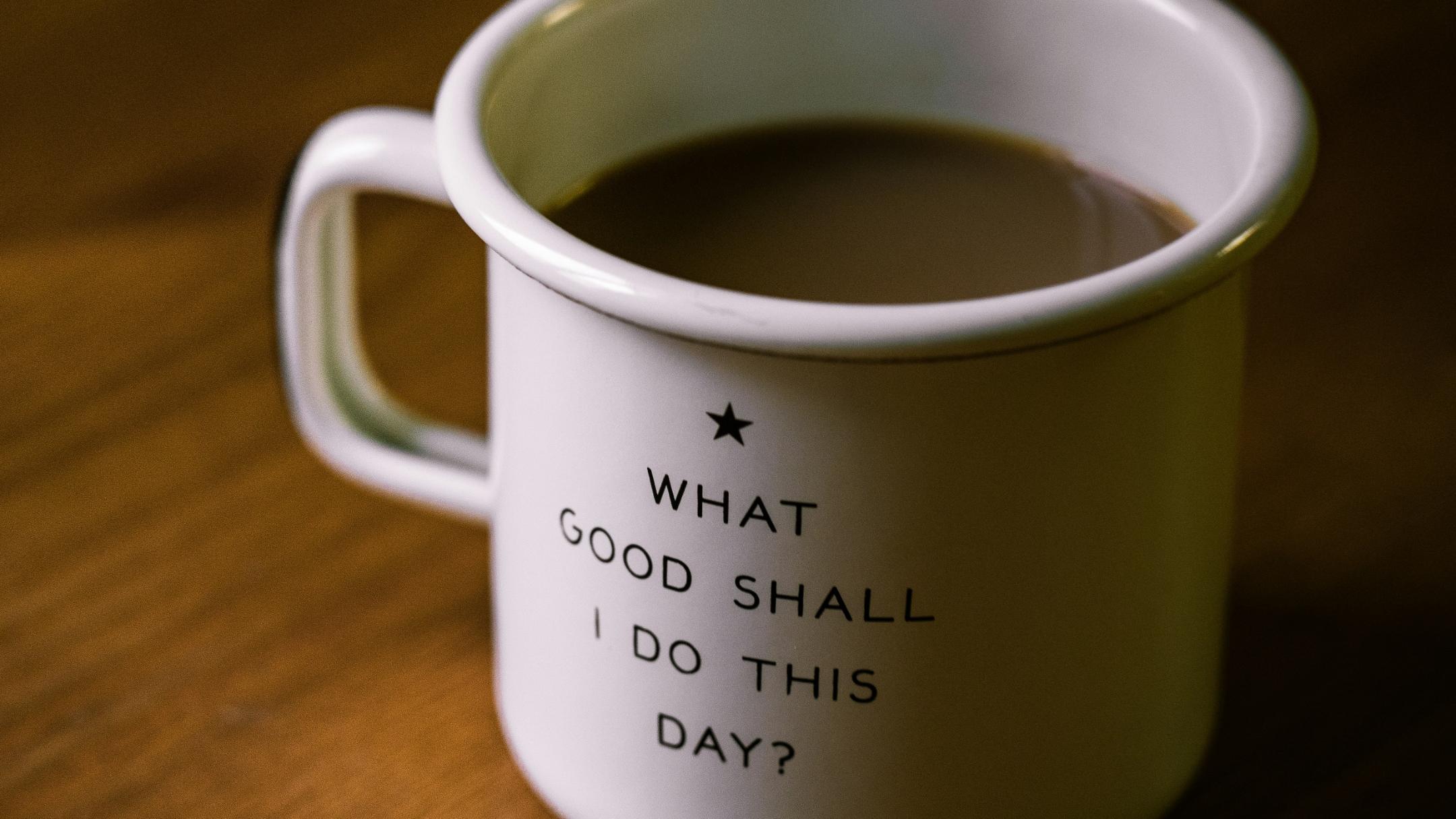 Weisse Tasse auf braunem Tisch; Aufschrift auf der Tasse: 'What Good Shall I Do This Day?'