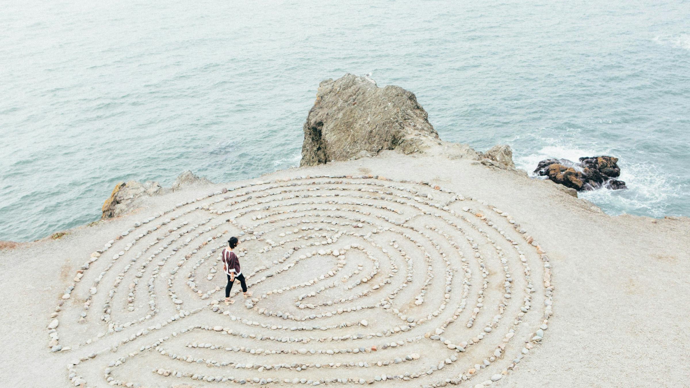 ein kreisrundes Labyrinth aus hellen Steinen auf hellem Sand direkt an der Küste, zwei kleinere Felsen bilden die Grenze zwischen Wasser und Land, auf oder in dem Labyrinth geht eine junge Frau